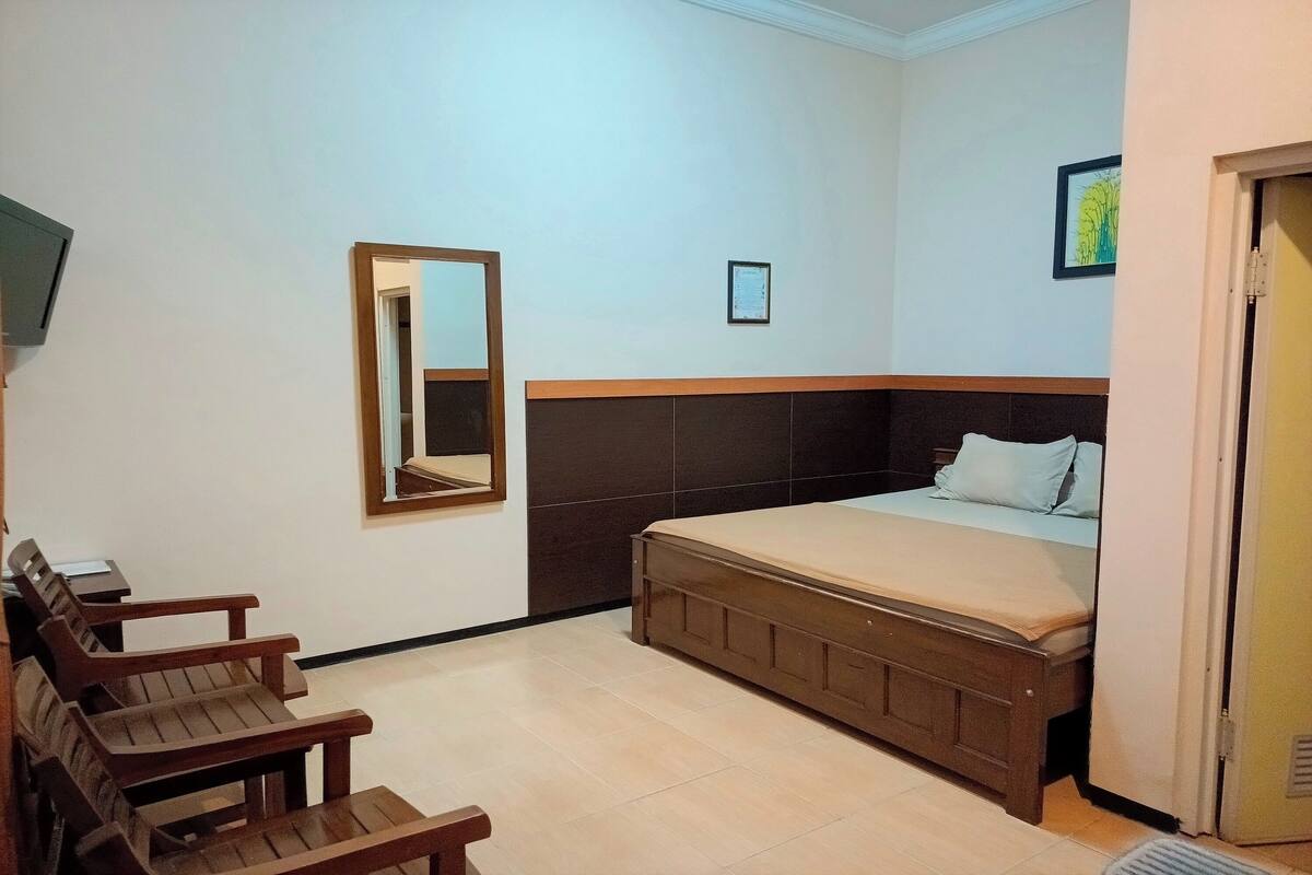 A hotel room with a bed, wooden chairs, a mirror, and a framed picture on the wall.