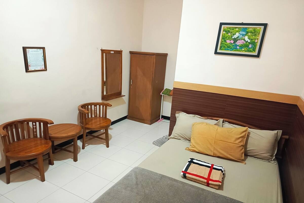 A bedroom with a bed, two wooden chairs, a small table, a wardrobe, and a framed picture on the wall.