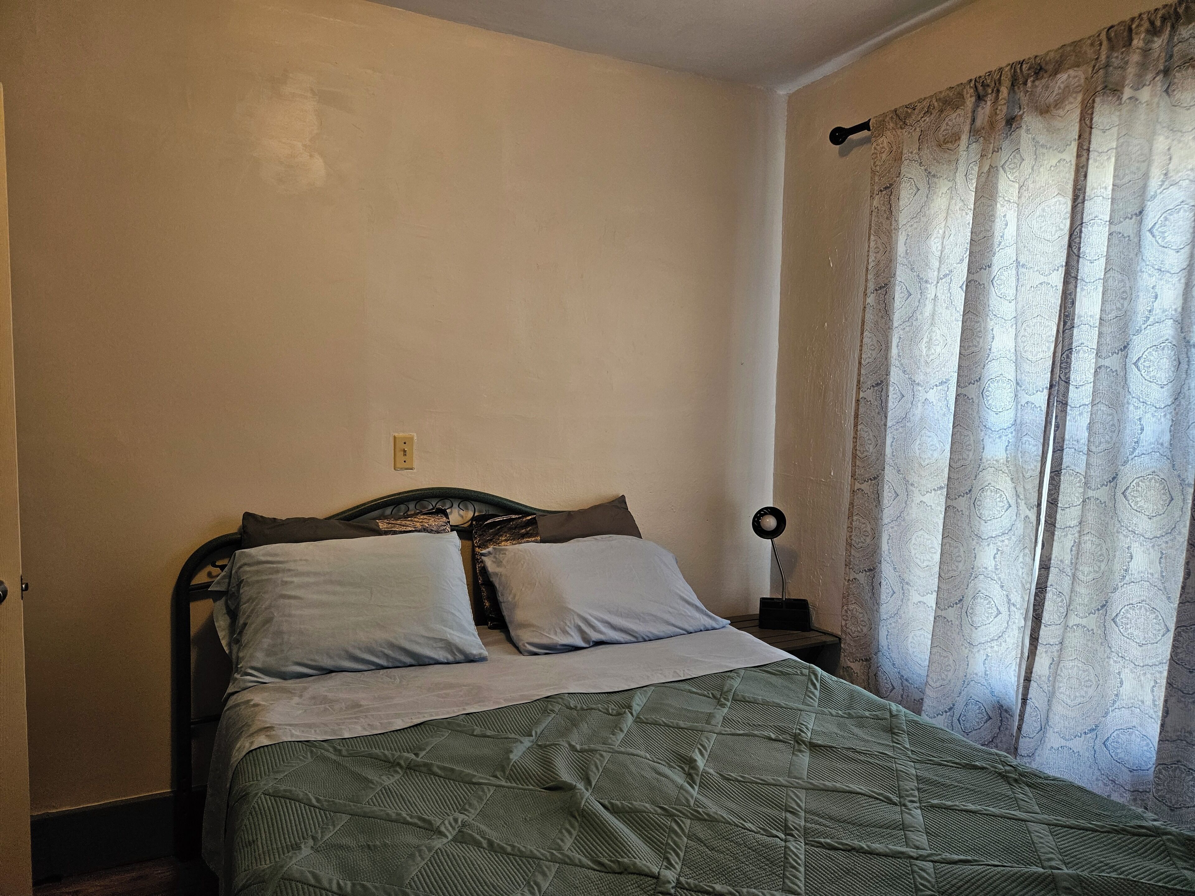 A single bed with a green quilt, two pillows, a bedside table with a lamp, and a window with patterned curtains.