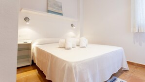 Una cama bien hecha con ropa de cama y toallas blancas, una mesita de noche con una lámpara, y un cuadro en la pared.