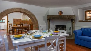 A dining area with a white table set for six, surrounded by white chairs. An archway leads to a kitchen area with a brick wall and a wooden table. A blue sofa is visible in the foreground. - Beautiful private villa with private pool, WIFI, terrace, pets allowed and panoramic view (Cortona)