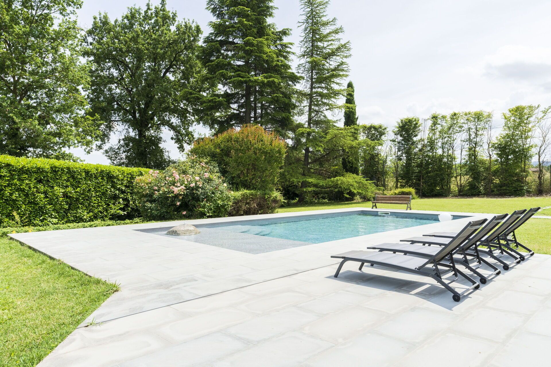A rectangular swimming pool with a stone accent in the center, surrounded by a paved deck. There are four lounge chairs aligned parallel to the pool, and a wooden bench is placed near the pool's edge. The area is bordered by a neatly trimmed hedge and various trees and shrubs.