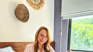 Une personne est assise sur un lit, avec un plateau rempli d’aliments pour le petit-déjeuner, notamment des œufs, du bacon et du jus d’orange.