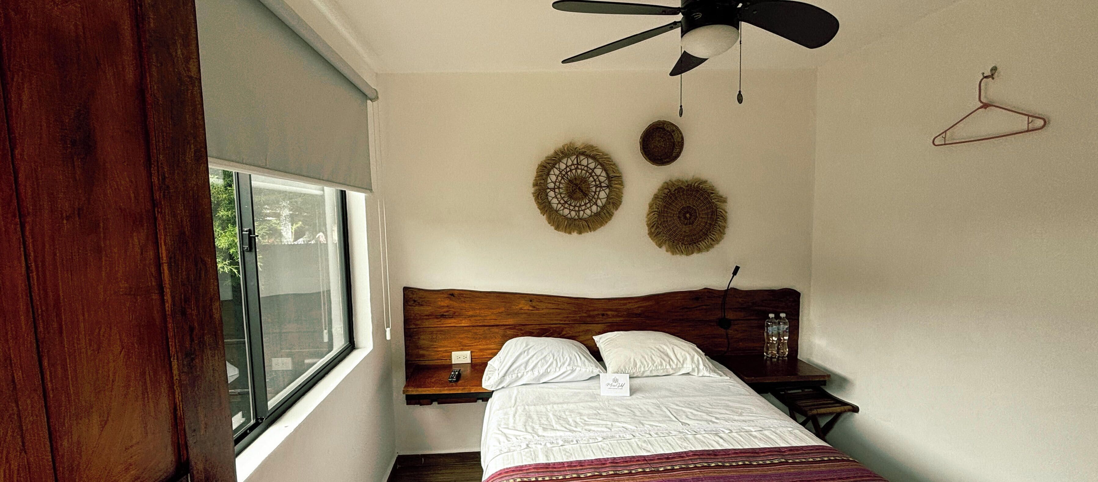 A bedroom with a wooden headboard, a bed with white bedding and a striped blanket, a bedside table with two bottles, a wall-mounted fan, and two wall hangings.