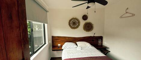 A bedroom with a wooden headboard, a bed with white bedding and a striped blanket, a bedside table with two bottles, a wall-mounted fan, and two wall hangings.