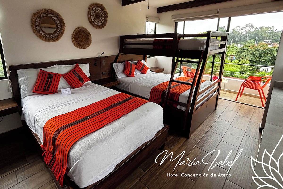 A hotel room with two bunk beds, a wooden nightstand, a small table, and a view of greenery through large windows.