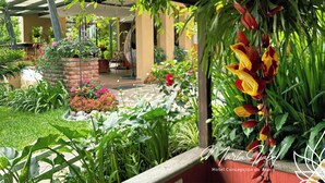 Un jardin luxuriant, avec une grande variété de plantes et de fleurs, un jardinier en briques et une pergola ornée de fleurs suspendues.