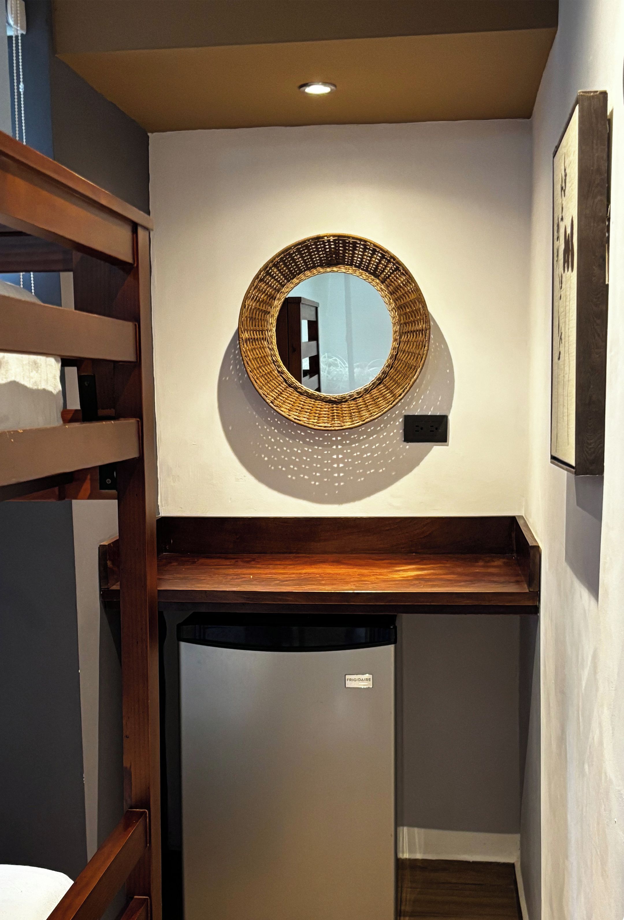 A compact kitchenette with a refrigerator, a wooden shelf, and a wall-mounted mirror.
