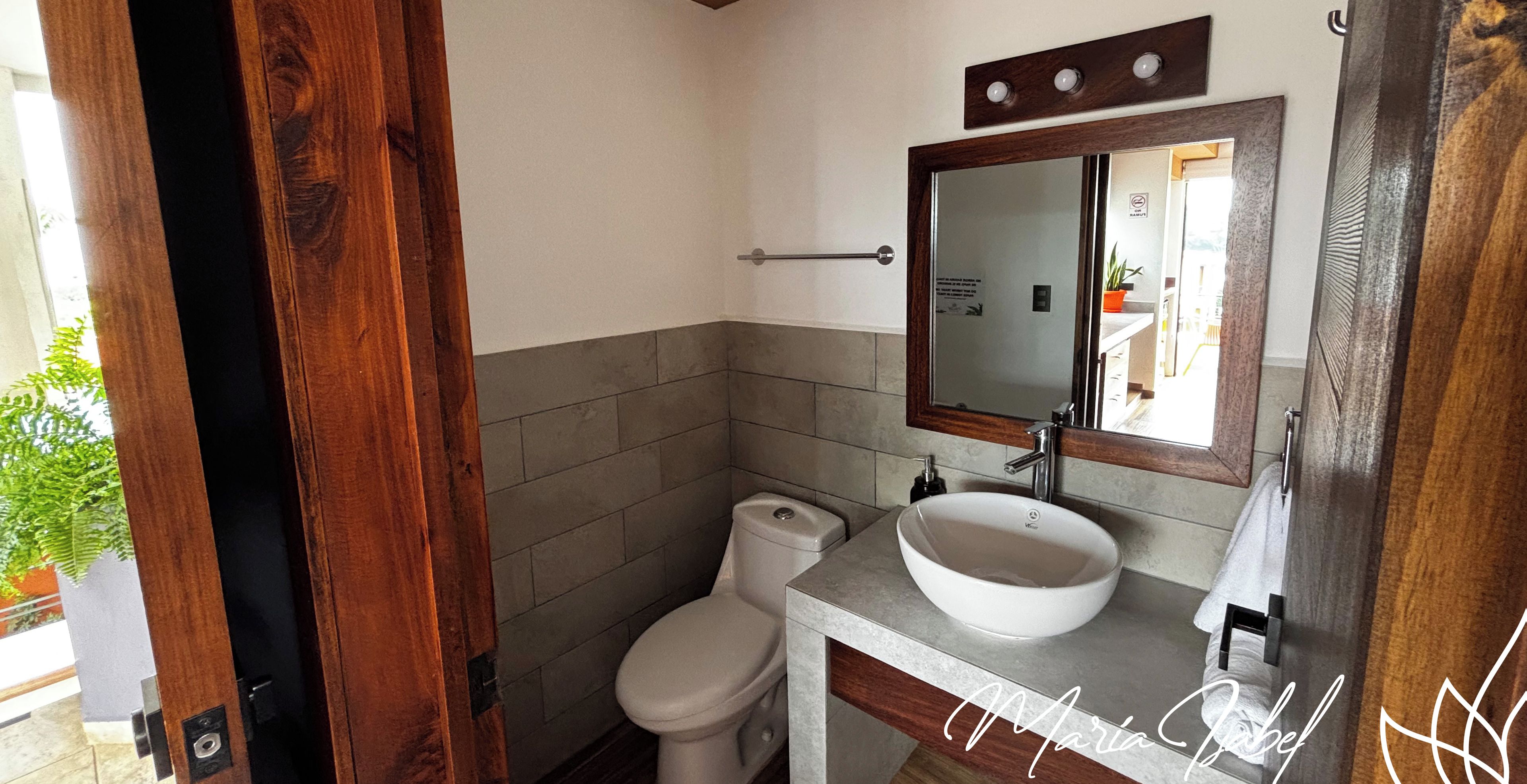 Un baño con puerta de madera, inodoro blanco, lavamanos con espejo y pared de azulejos.