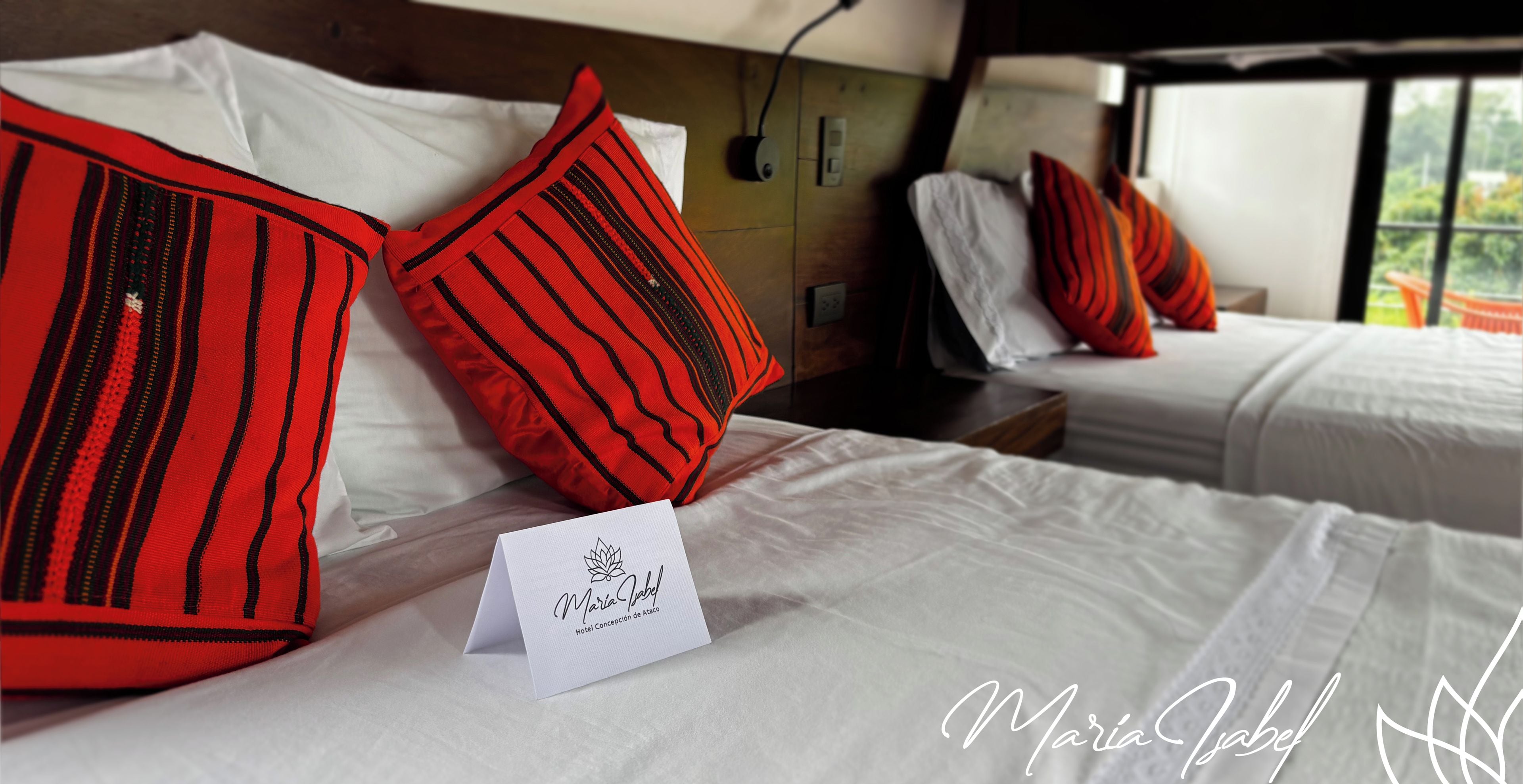 A hotel room with two beds, each with white bedding and red striped pillows. A white card with the hotel's name is placed on the bed.