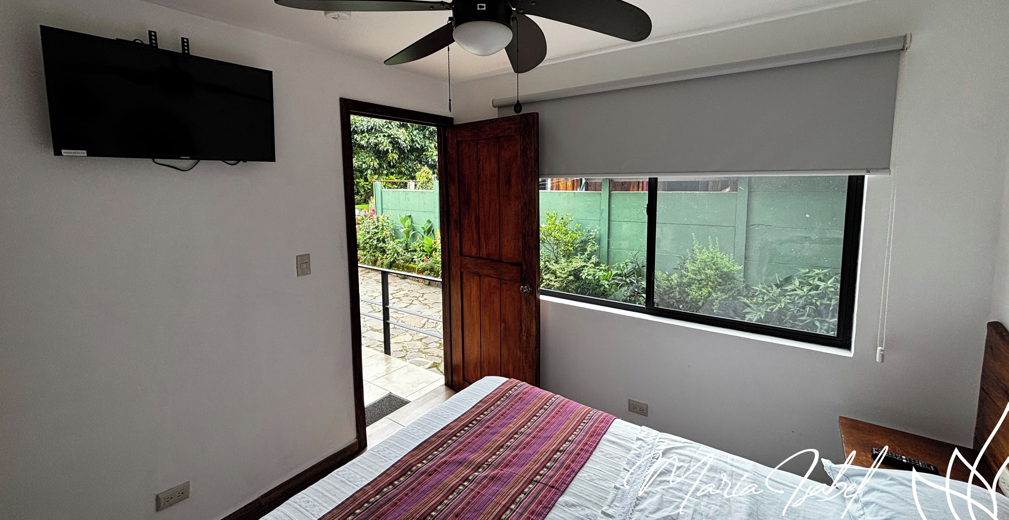 Un dormitorio con cama, televisor montado en la pared, ventilador de techo y ventana con vistas a la vegetación.