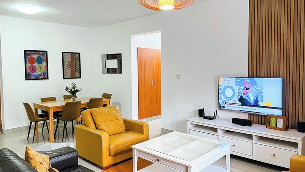 A modern living room with a sofa, coffee table, yellow armchair, dining table, chairs, and a television. - 3 Bed Apartment - Sleeps 6 - Free Parking (Luanda)
