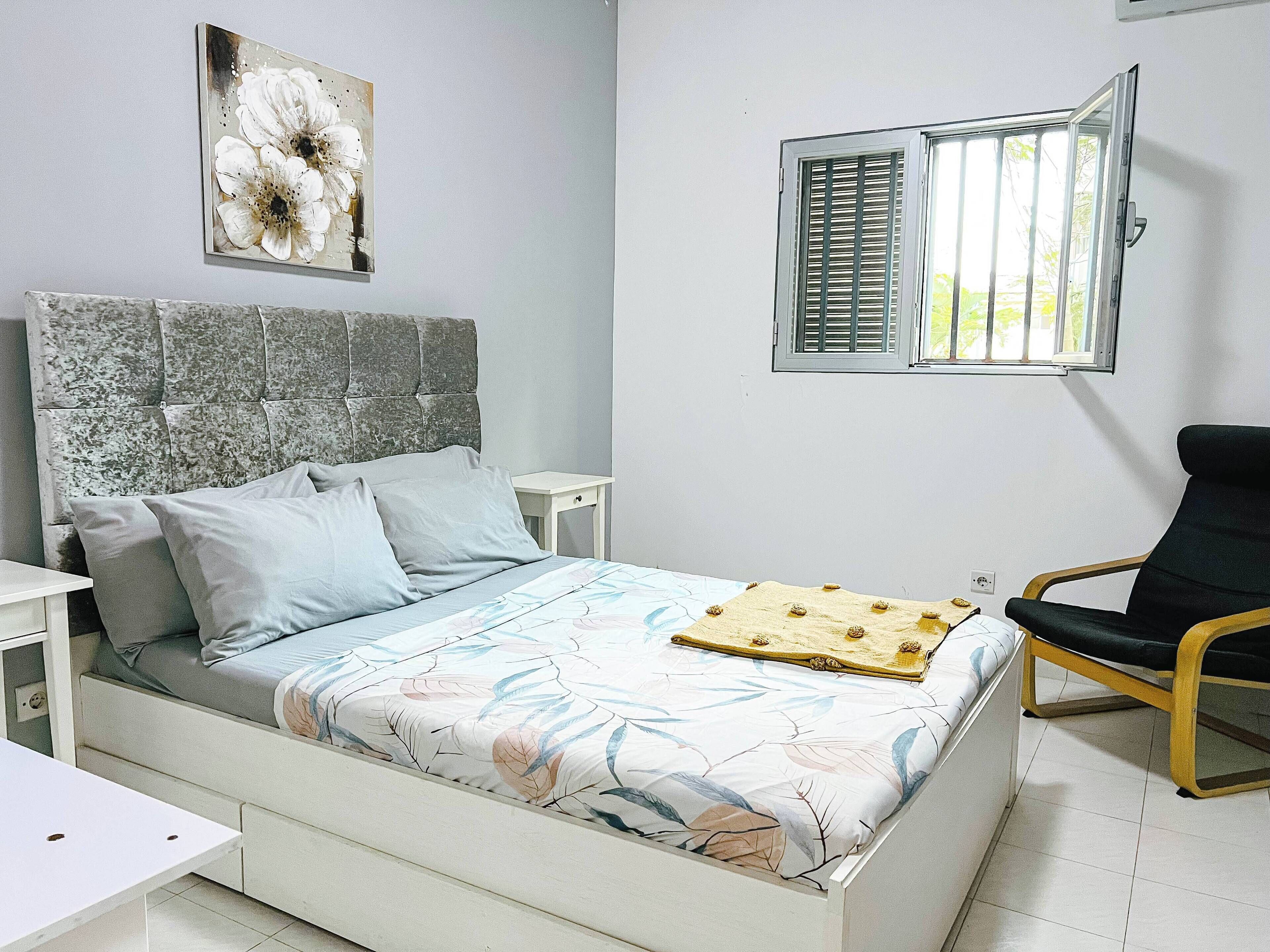 A bedroom with a bed, a nightstand, a chair, a window with shutters, and a wall-mounted artwork.