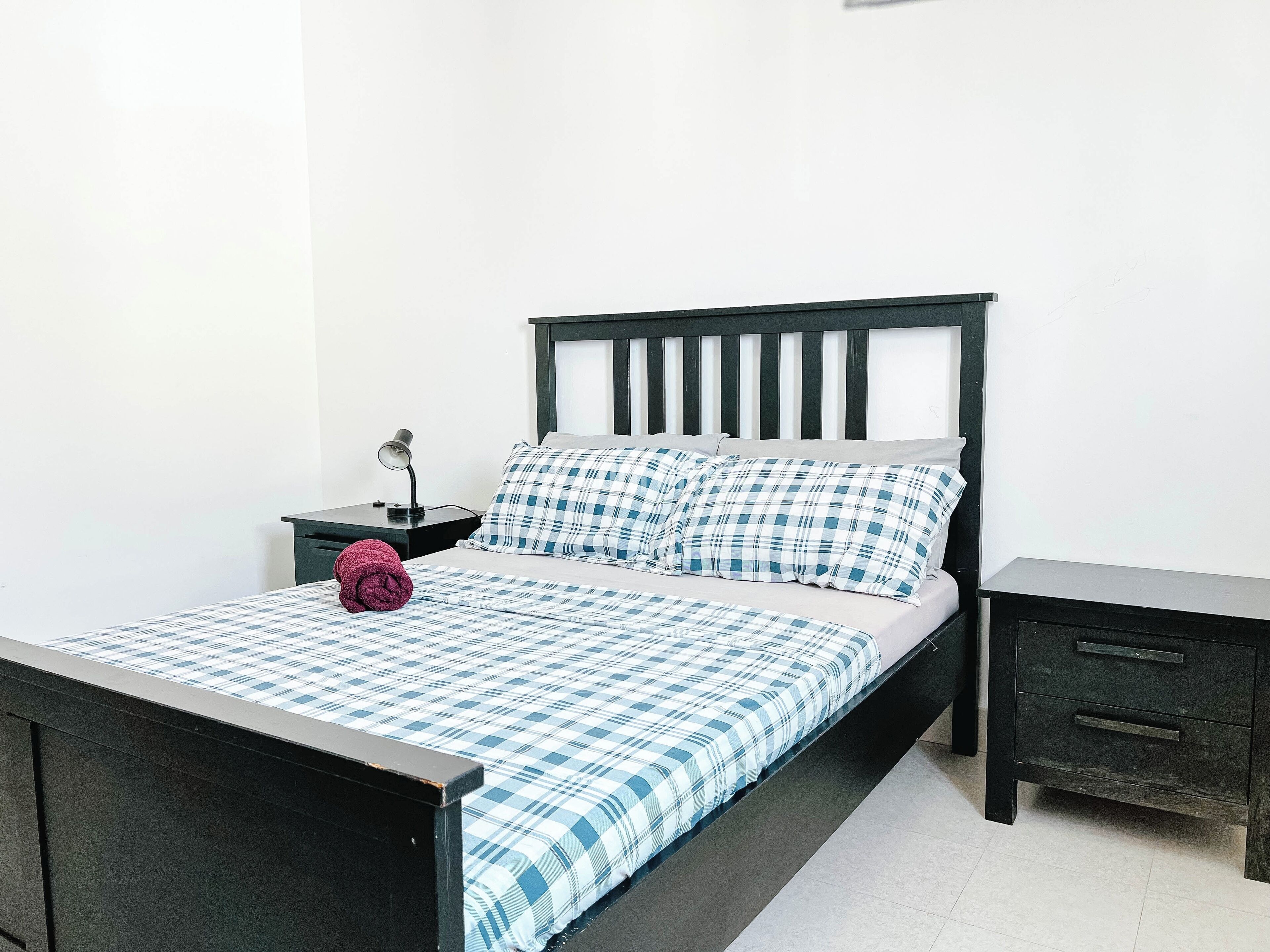 A bed with a checkered bedspread, a pillow, a nightstand with a lamp, and a drawer unit.