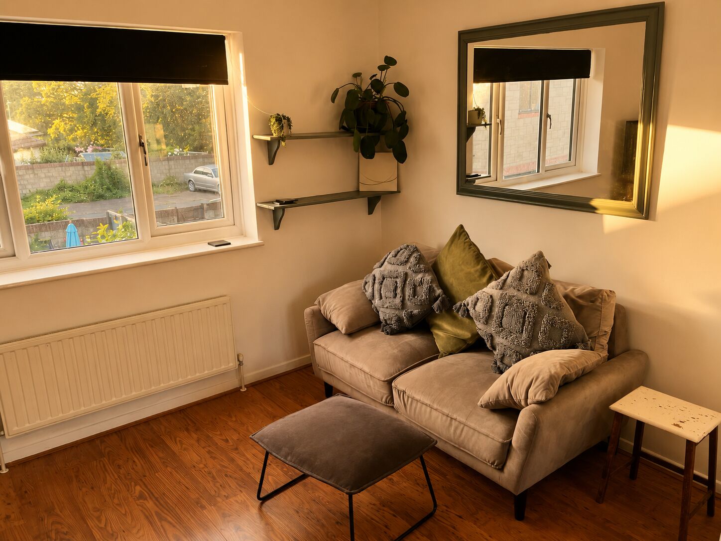 A room with a beige sofa, a small wooden stool, a radiator, a window with blinds, and a mirror on the wall.