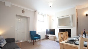 A living room with a grey sofa, a blue armchair, a TV, a fireplace, and a dining table set for four. - Haven 3 Bedroom House in Whalley (Clitheroe)