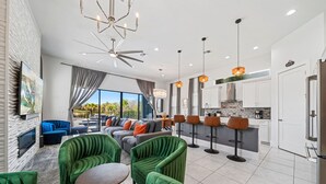 A modern living room with green upholstered chairs, a sofa, and a flat-screen TV. There is a kitchen area with white cabinets and a dining bar with stools. - Fairway Prosperity: Arcade, Themed Bedroom, Golf, Private Pool, Near Disney (Kissimmee)
