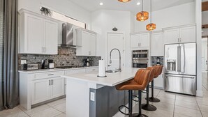 A modern kitchen with white cabinetry, a central island with bar stools, stainless steel appliances, and a brick backsplash. - Fairway Prosperity: Arcade, Themed Bedroom, Golf, Private Pool, Near Disney (Kissimmee)