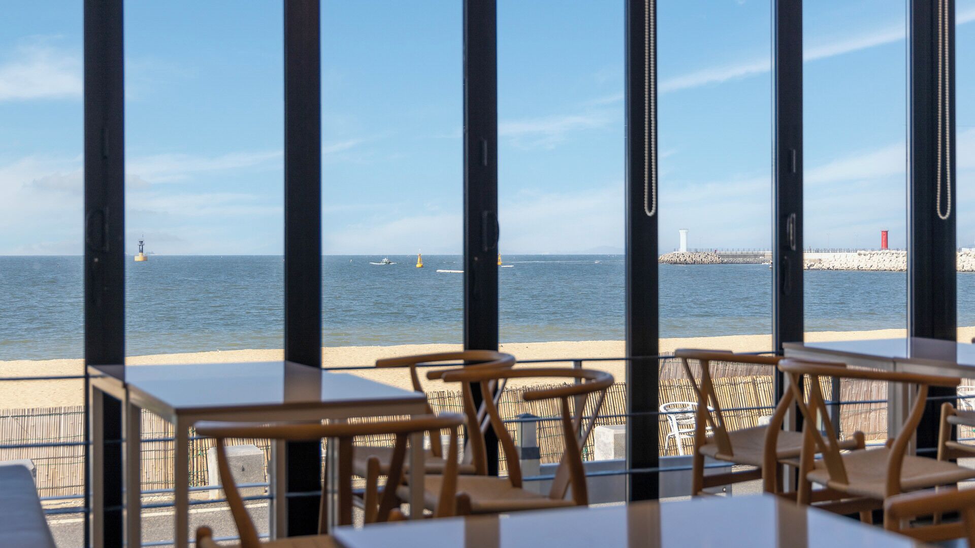 Um restaurante à beira-mar com mesas e cadeiras voltadas para o oceano, um píer com um farol e uma bóia vermelha.