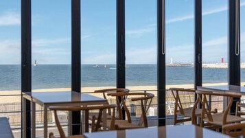 A seaside restaurant with tables and chairs facing the ocean, a pier with a lighthouse, and a red buoy.