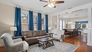 A spacious living room with a brown leather sofa, a grey armchair, and a wooden coffee table. A dining area with a wooden table and chairs is visible in the background. - Maple Haus Entire Property | Sleeps 18 | Great Location (Fredericksburg)