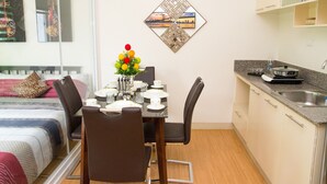 Una habitación de hotel compacta con una mesa de comedor lista para dos personas, una pequeña cocina y una cama con un cojín estampado.
