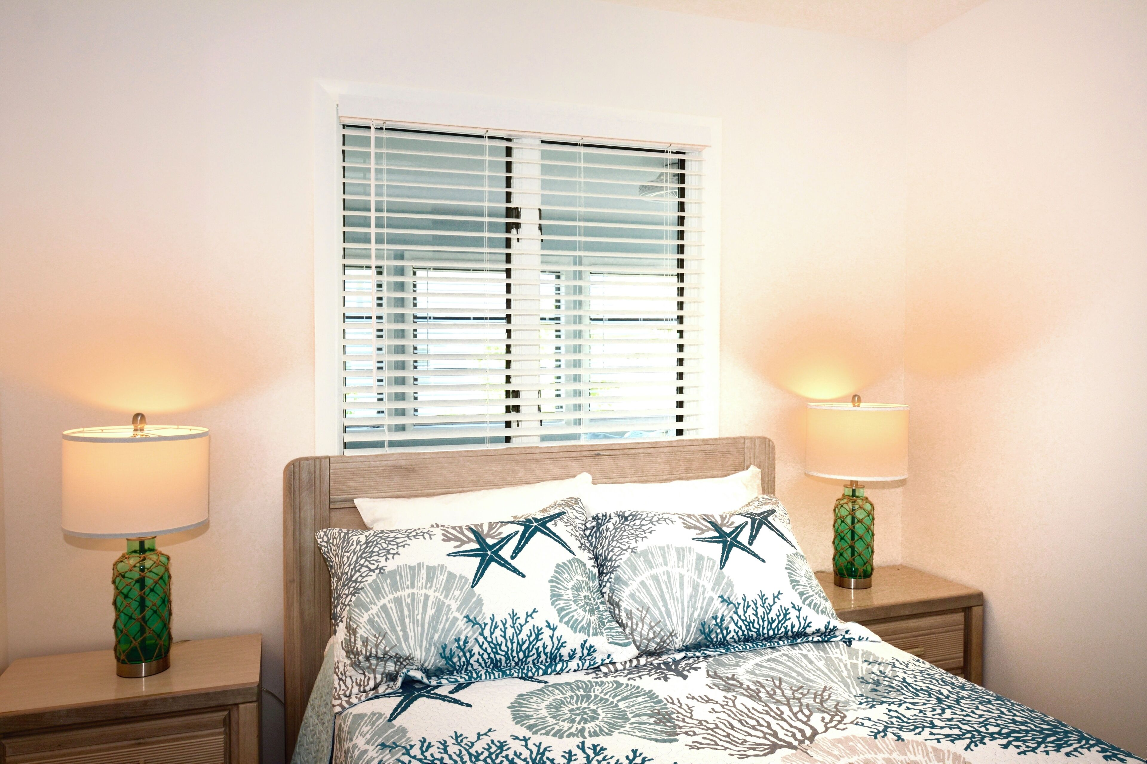 A bedroom with a bed, two bedside lamps, a window with blinds, and a nightstand.