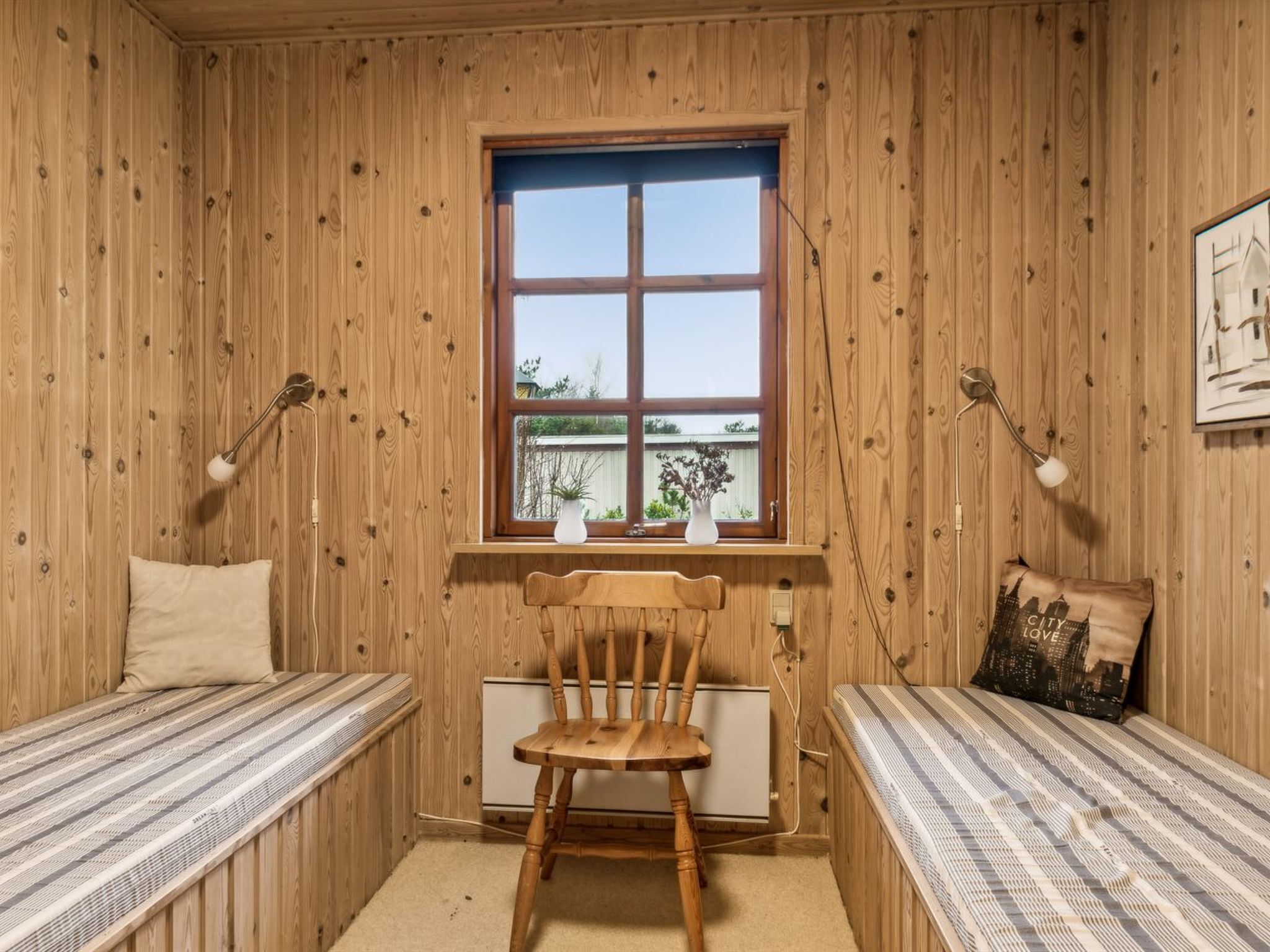 Una habitación con paredes de madera, un ventanal grande, dos mesitas de noche con lámparas, una silla y una cama.