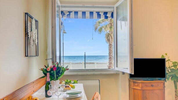A dining area with a table set for two, a television, and a window with a view of the sea. - Mer Vivante - Front Beach with Balcony, Albenga, Italy (Albenga)