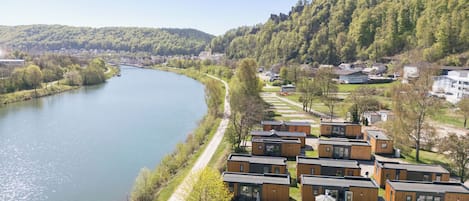 A riverside view with wooden cabins, a clear sky, and a forested hill.