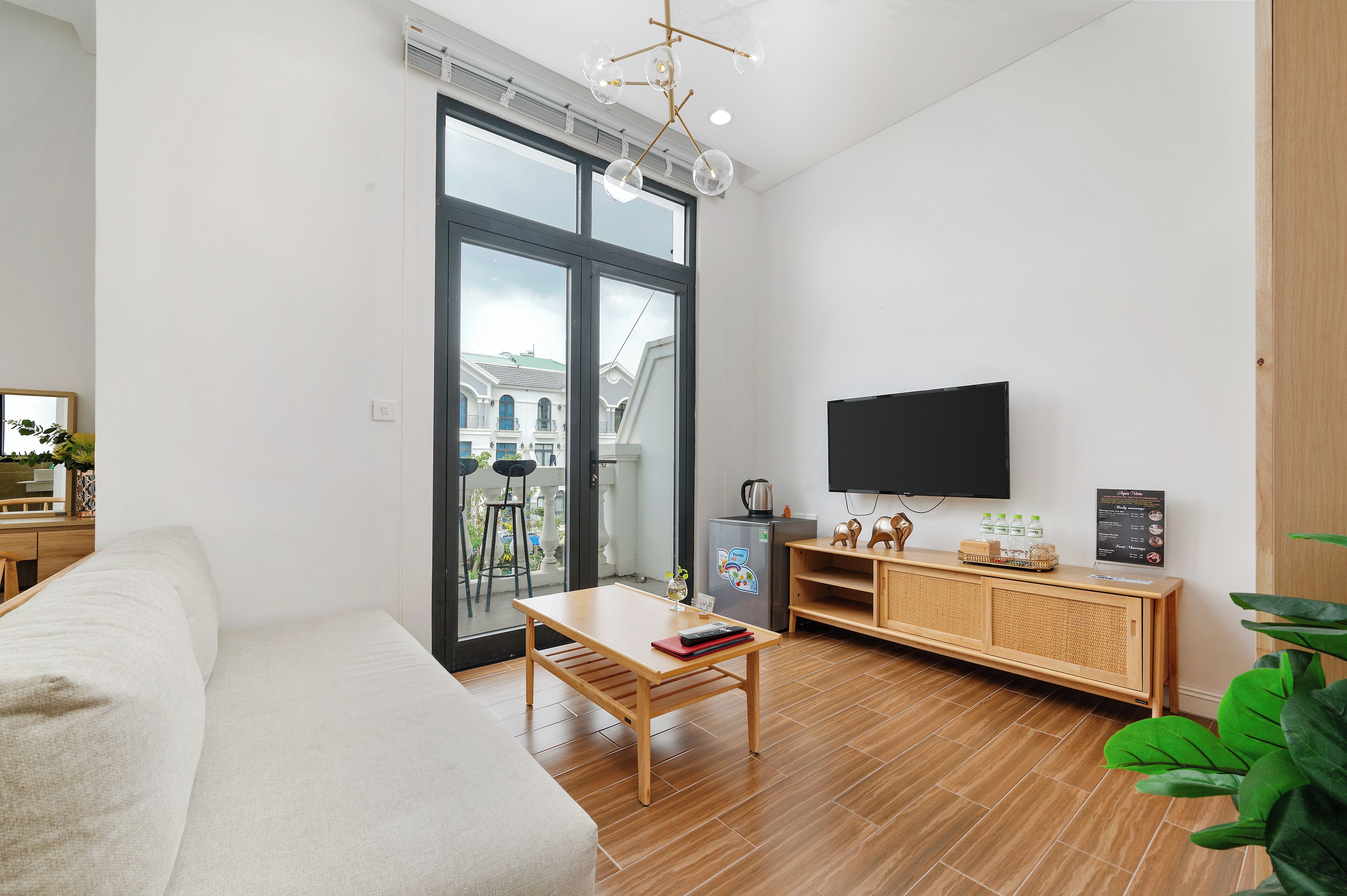 A modern living room with a sofa, a wooden coffee table, a TV mounted on the wall, and a large window offering a view of the outside.