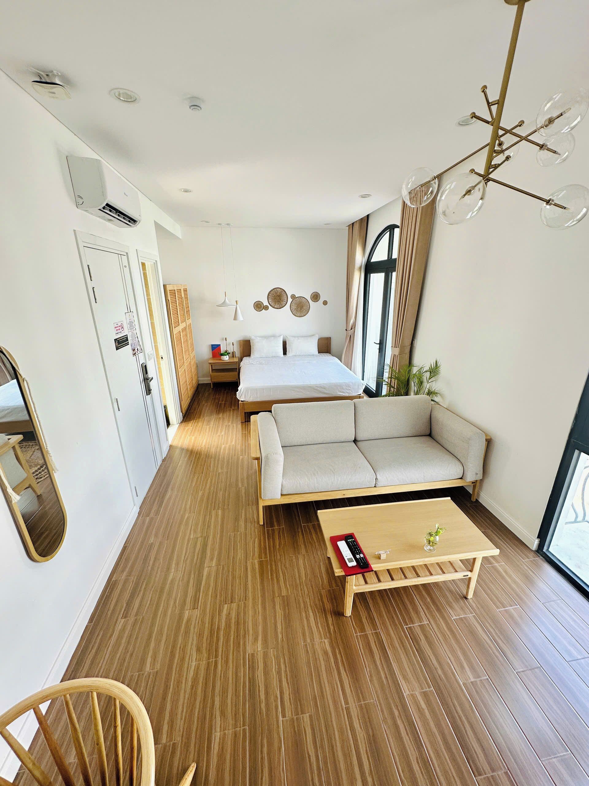A modern hotel room with a bed, sofa, and a small table. There is a mirror, a window, and a ceiling light fixture.