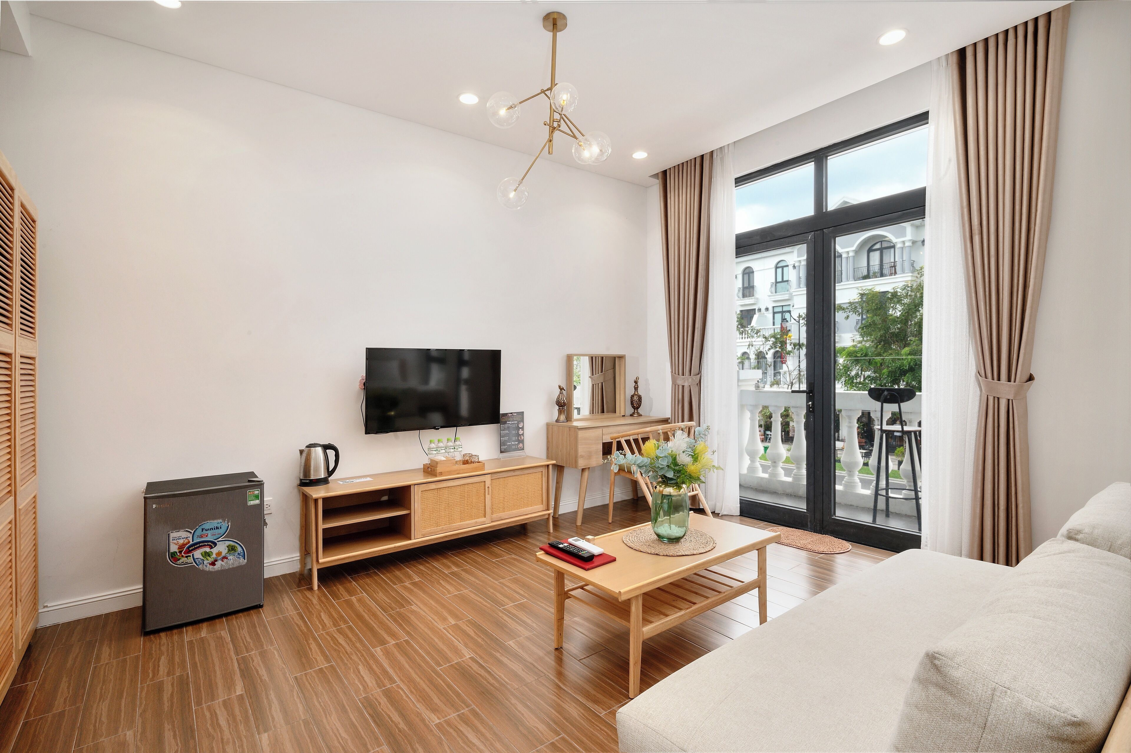 A modern living room with a flat-screen TV, wooden furniture, a small refrigerator, and a large window offering a view of buildings and greenery.
