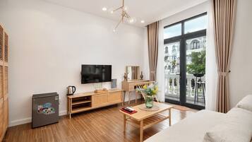 A modern living room with a flat-screen TV, wooden furniture, a small refrigerator, and a large window offering a view of buildings and greenery.