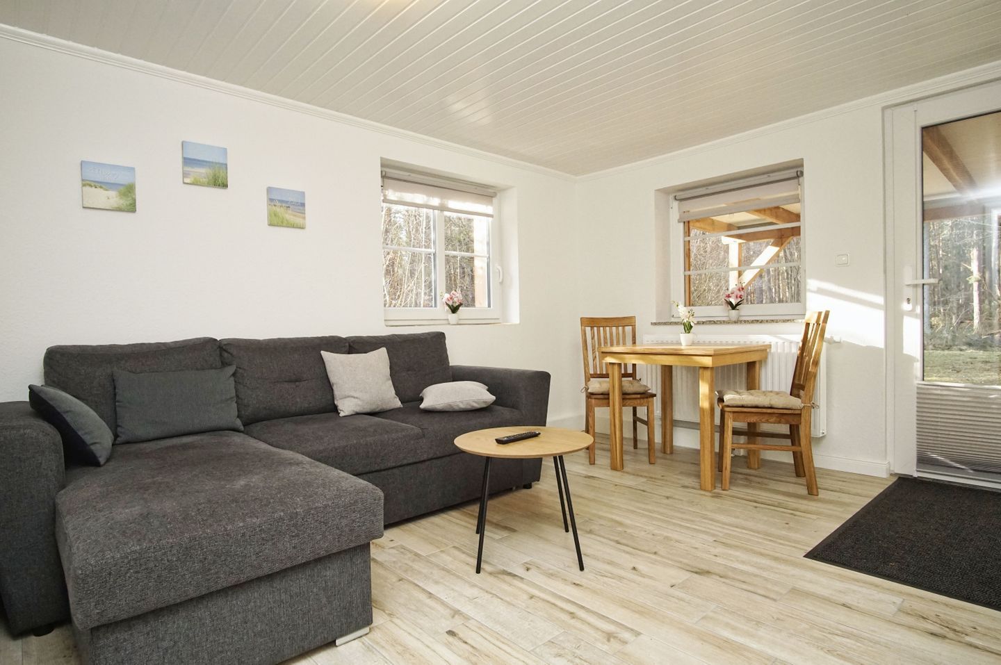 Een moderne woonkamer met een grijze hoekbank, een klein rond tafeltje en een houten eettafel met stoelen. Er zijn twee ramen met zonwering, een radiator en een deur naar buiten.