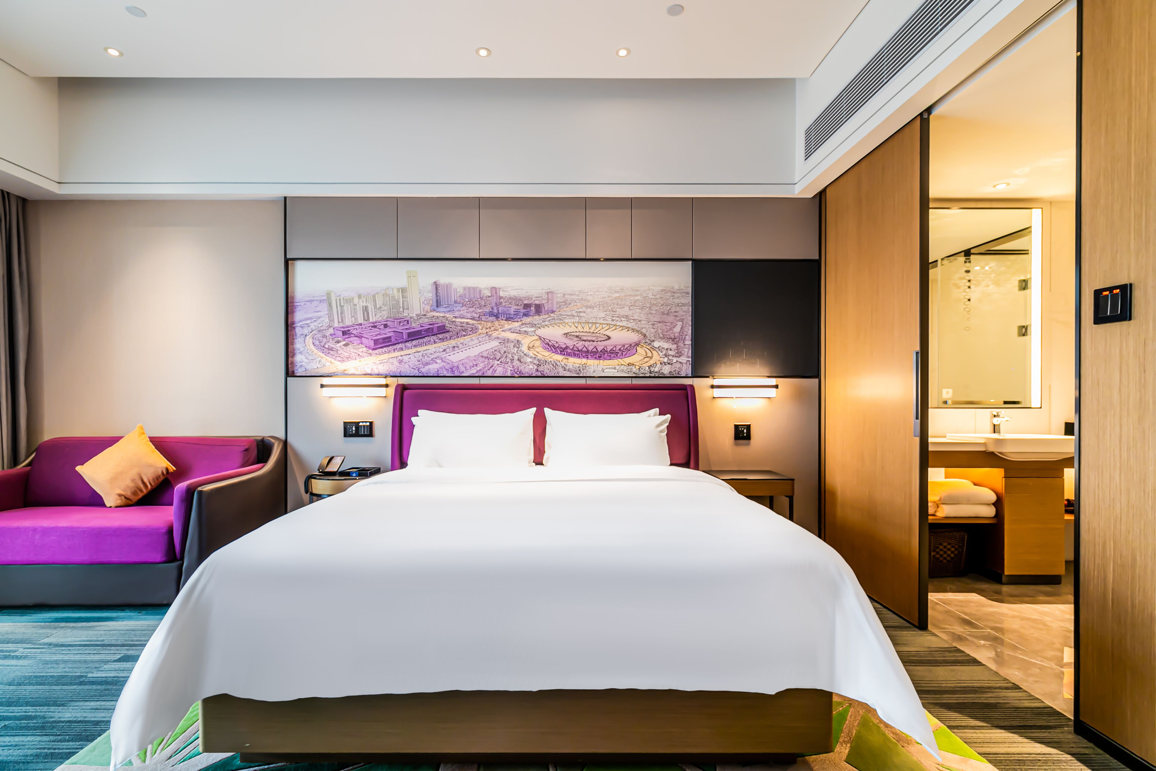 A hotel room with a large bed, a purple sofa, a cityscape artwork on the wall, and a bathroom visible through an open door.