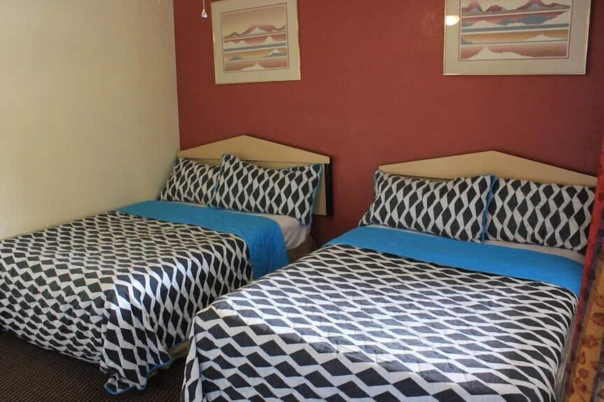 A room with two beds, each with patterned bedding and a blue blanket. There are two framed pictures on the wall.