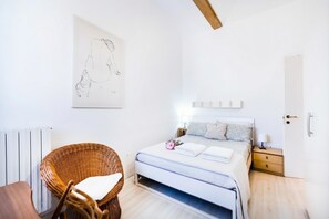 Quarto com cama, cadeira de vime, mesa de cabeceira com luminária e um quadro na parede.