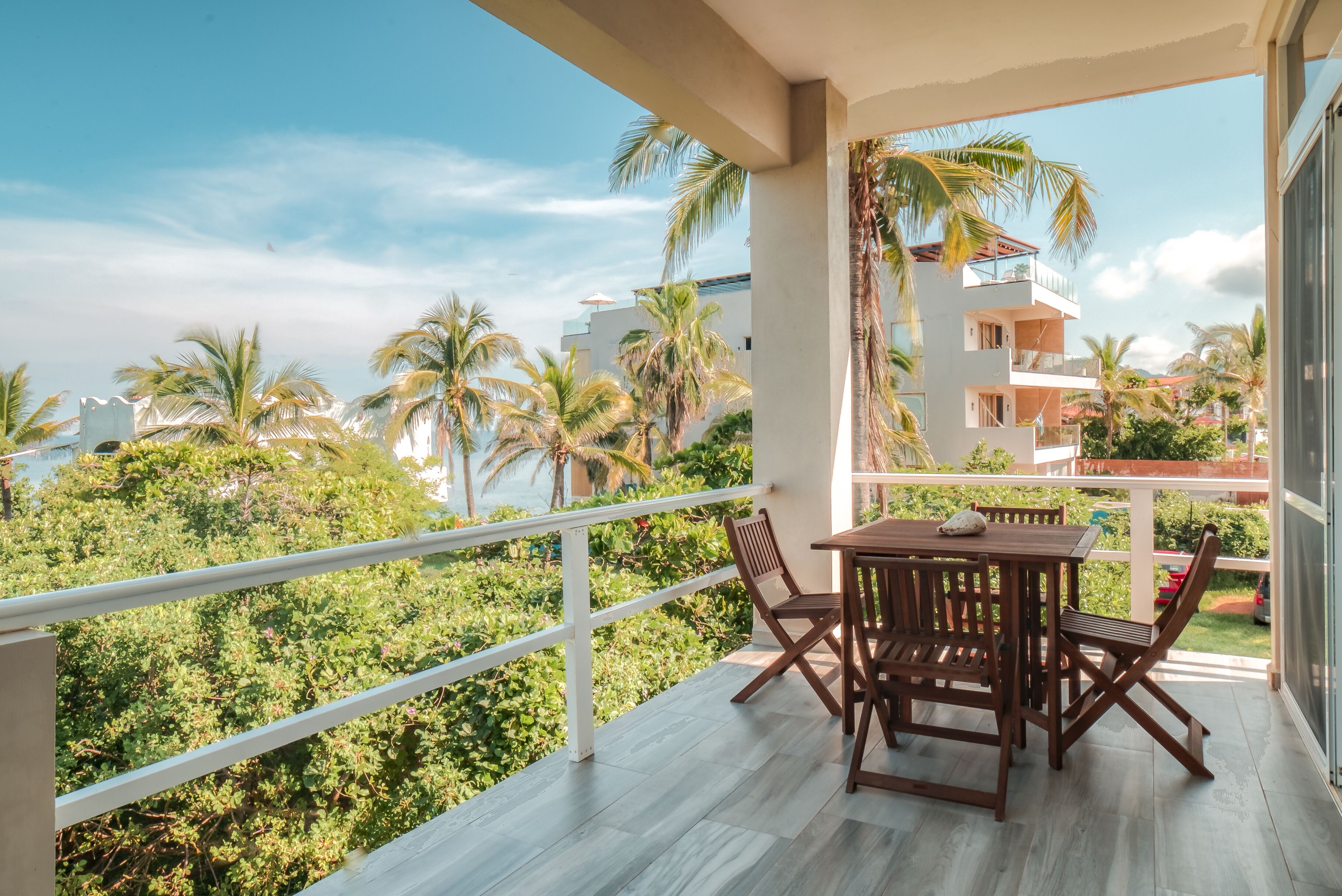 A balcony with a wooden table and chairs, overlooking a lush green landscape and a white building with balconies.