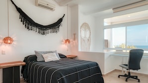 Un dormitorio con cama, mesitas de noche, escritorio, silla, hamaca, decoración de macramé en la pared y un ventanal con vistas.