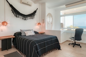 Un dormitorio con cama, mesitas de noche, escritorio, silla, hamaca, decoración de macramé en la pared y un ventanal con vistas.