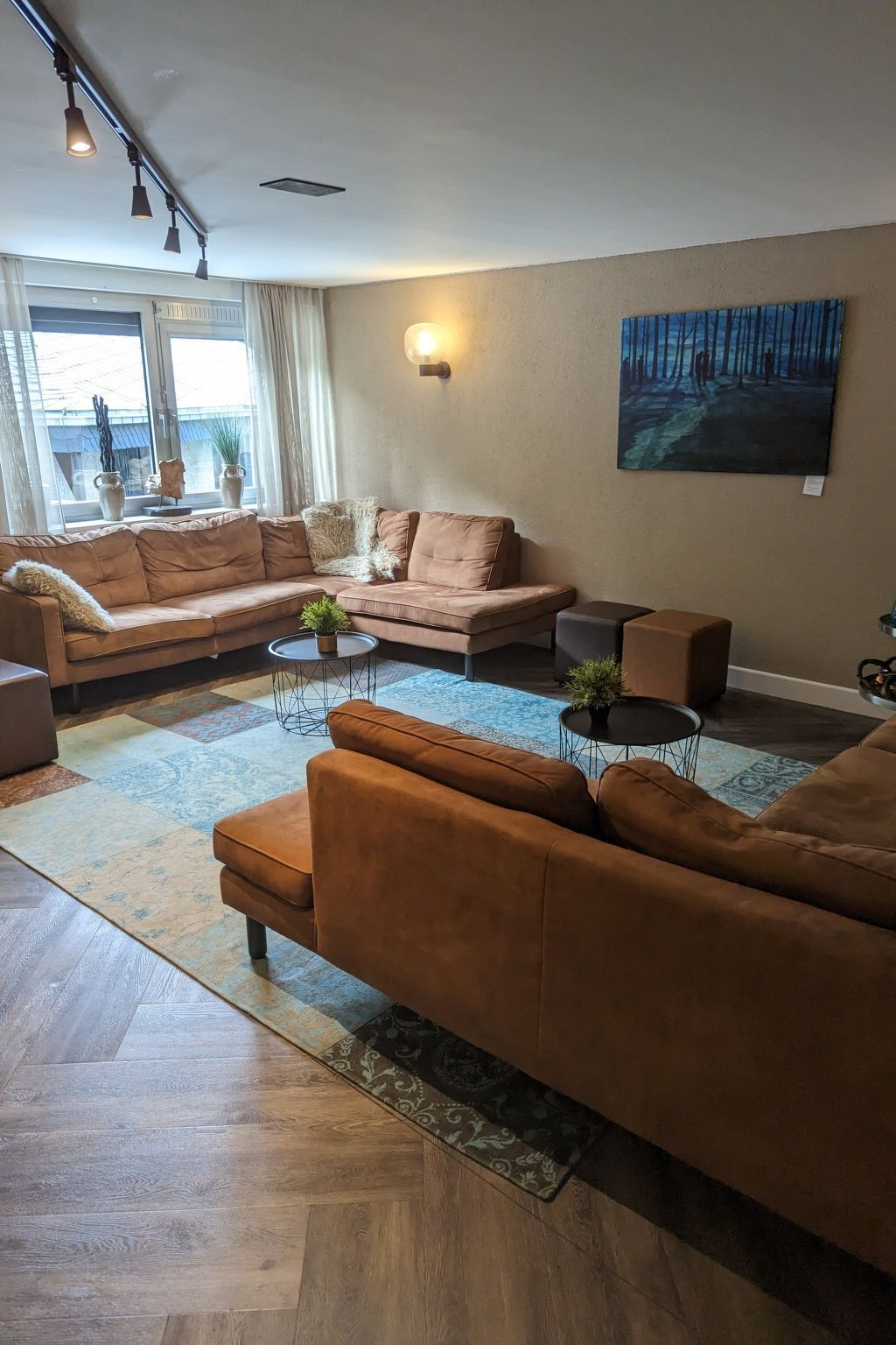 A modern living room with a large sectional sofa, a smaller sofa, a coffee table, and a side table. There is a painting on the wall, a window with curtains, and a floor lamp.