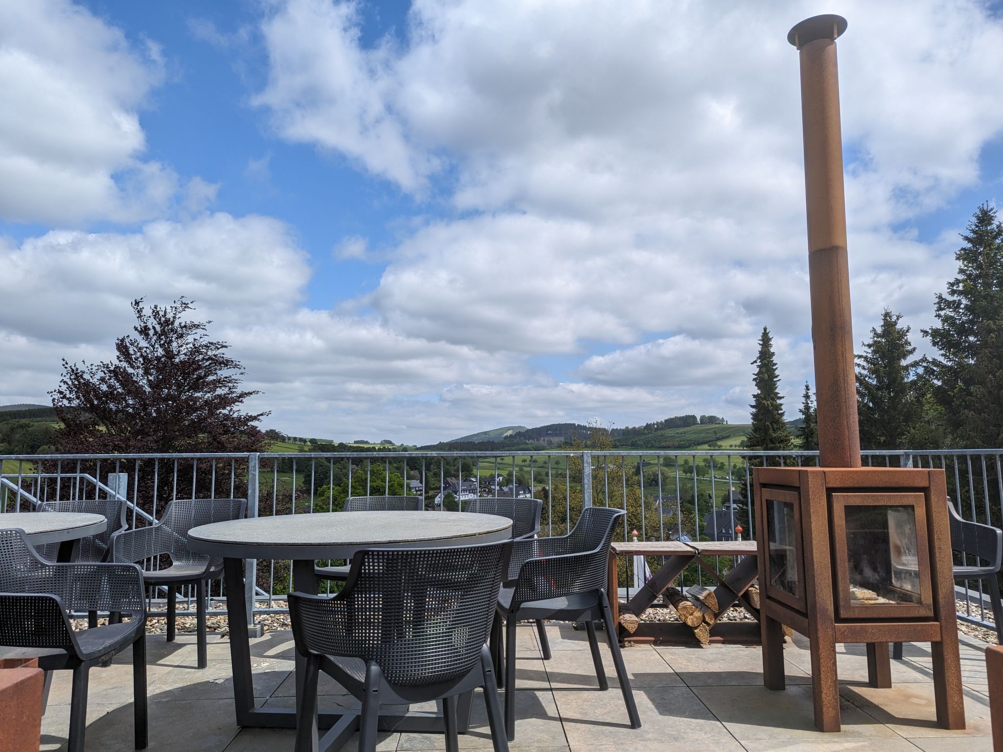 Een terras buiten met ronde tafels en stoelen, een houtkachel en een mooi uitzicht op heuvels en bomen.