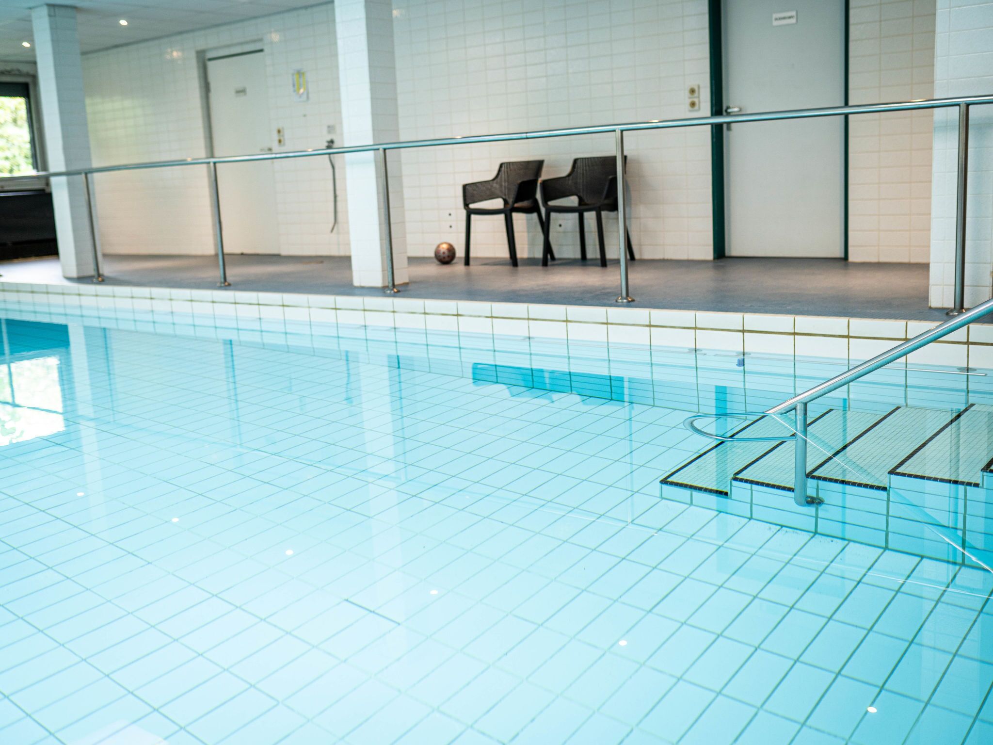 An indoor swimming pool with tiled flooring, a glass railing, and two black chairs against the wall.