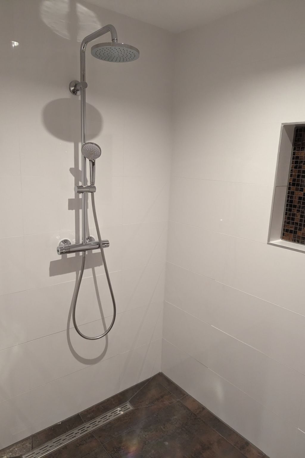 A shower with a handheld showerhead and a wall-mounted rain showerhead in a bathroom with white tiled walls and a small window with a decorative grille.