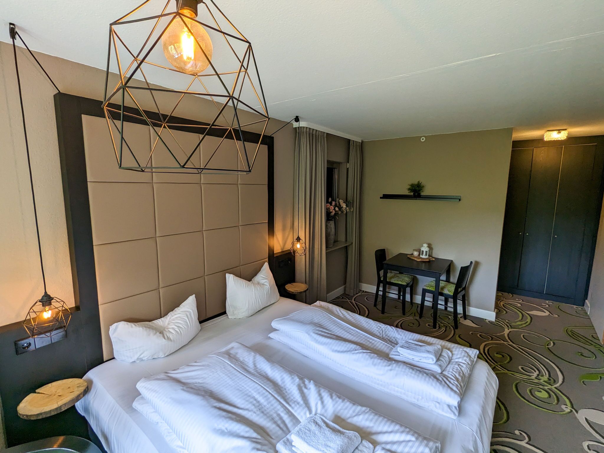 A hotel room with a large bed, a desk with chairs, a small table with a vase of flowers, and a wardrobe.