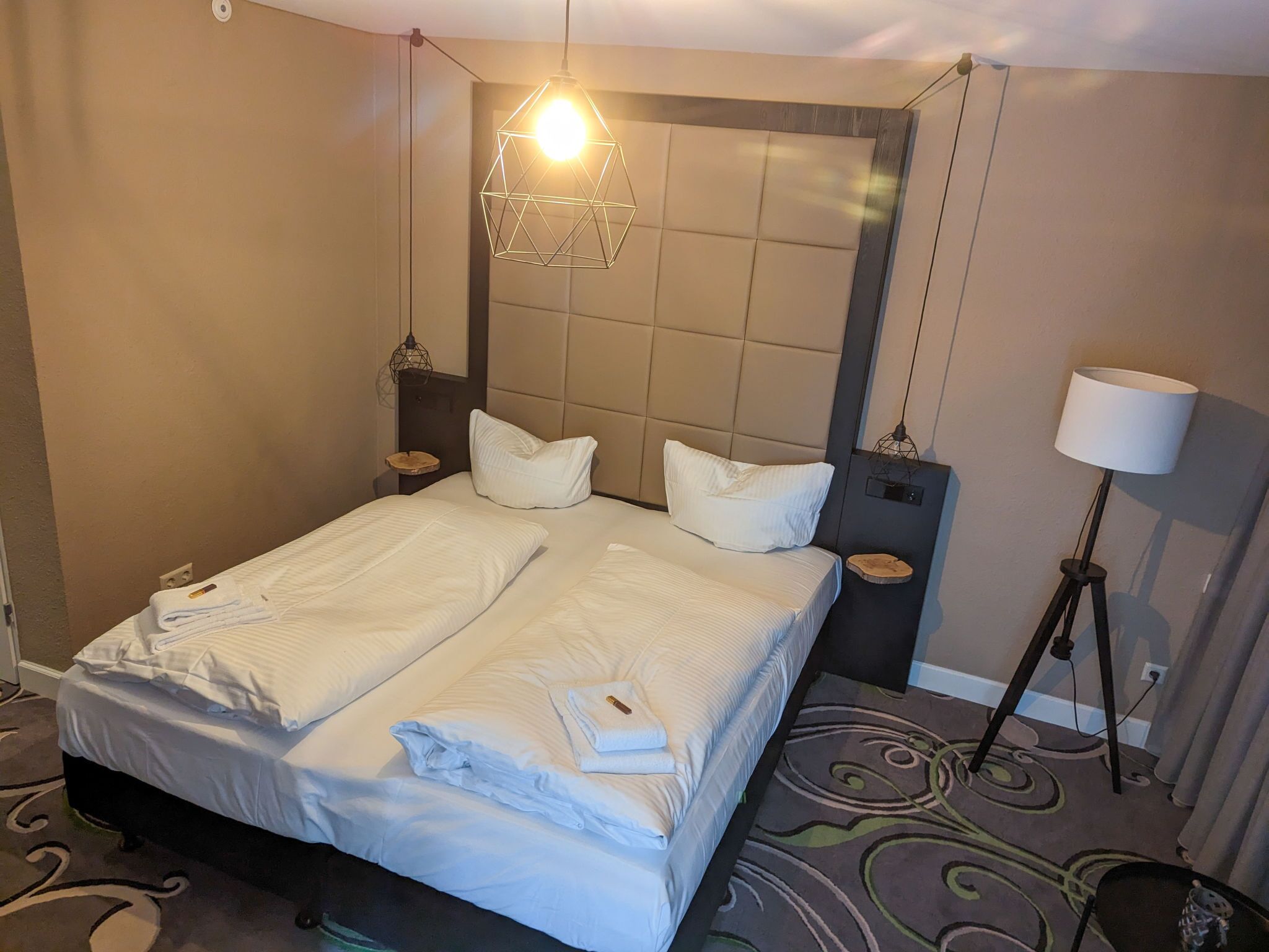 A hotel room with a double bed, two bedside tables, a floor lamp, and a geometric light fixture.