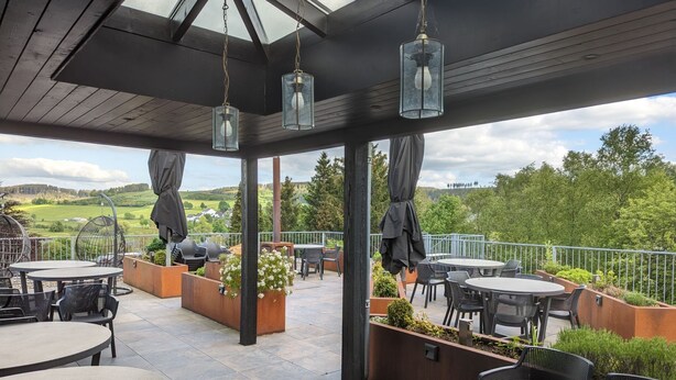An outdoor patio with round tables and chairs, a wooden ceiling, and a view of green hills. - Gruppe Meisenberg by Interhome (Winterberg)