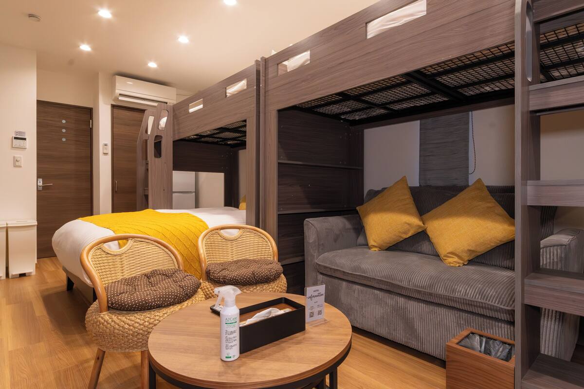 A hotel room with a bed, a sofa, a wicker chair, a small table, and an air conditioning unit.