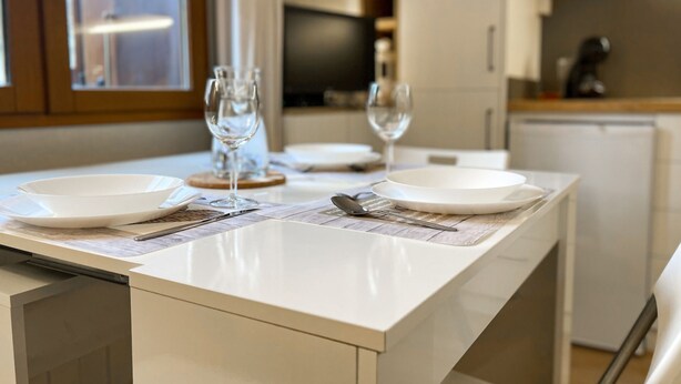 Une cuisine moderne avec des meubles blancs, un plan de travail en marbre et un coin repas agrémenté d’assiettes blanches, de couverts et de verres à vin.